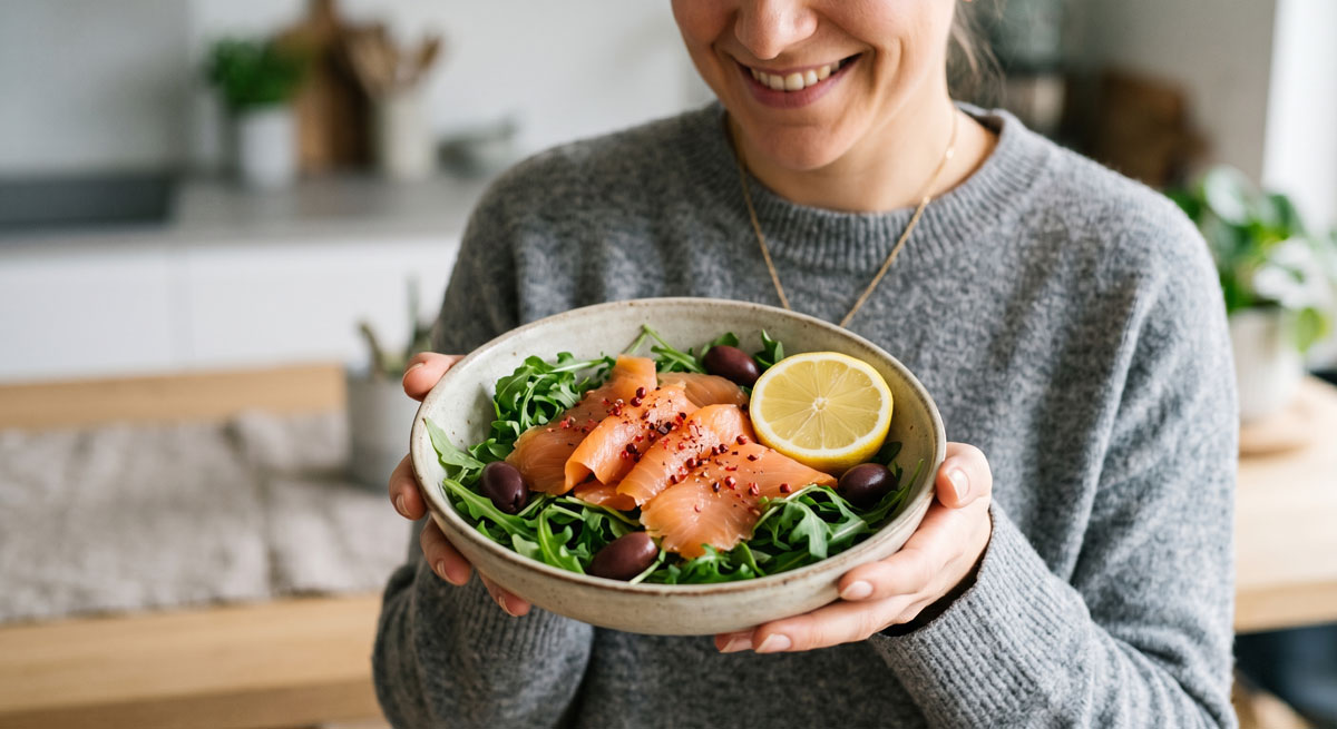 Person in a gray sweater holding a bowl of salad with smoked salmon, olives, arugula, and a lemon wedge in a kitchen.